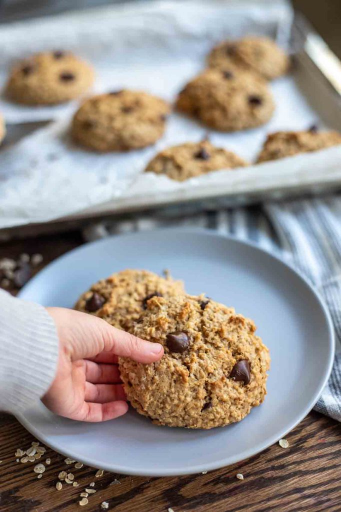 child's hand picking up a healthy sourdough cookie off of a gray plate with more cookies in the background on a baking sheet