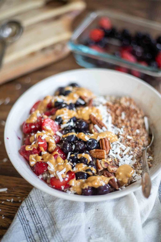 close up picture of a bowl of steel cut oatmeal topped with fruit, coconut, chia seeds, and drizzled with peanut butter with an antique spoon in the oatmeal. The bowl rests on white and blue stripped napkin on a wooden table with berries in a dish in the background