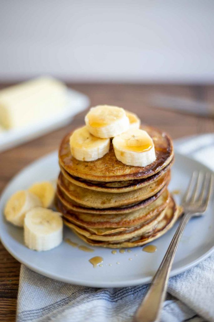 gluten free banana pancakes stacked up on a gray plate and topped with more sliced bananas. the plate sits on a white and blue napkin and a butter dish is seen in the background