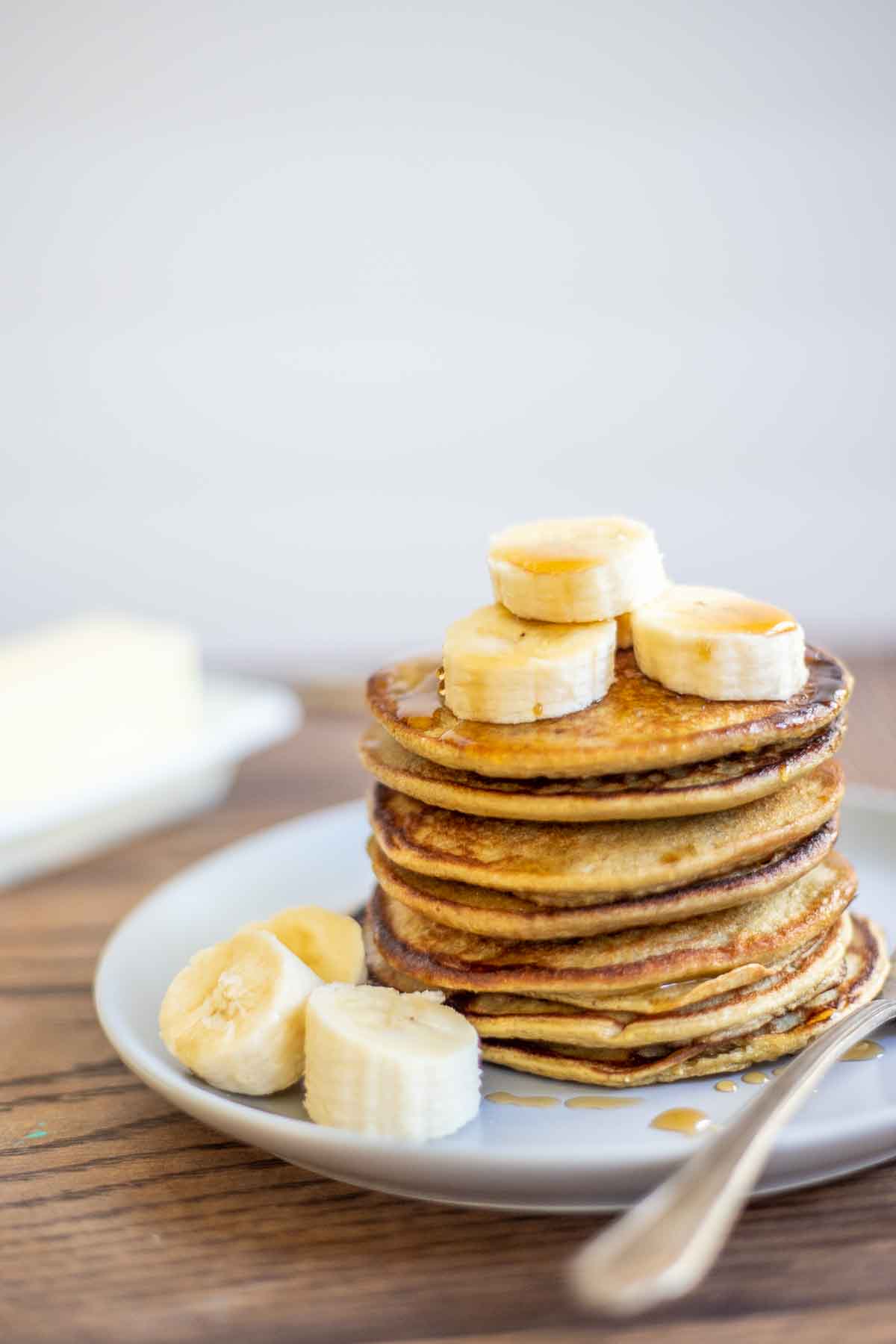 Stack of gluten free high protein banana pancakes with sliced banana on top. Pancakes are on a plate with a fork lying on the right side. There is a stick of butter in the background.