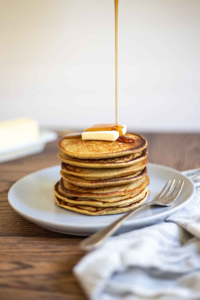 Stack of protein banana cakes on plate with syrup being poured on top. Towel in front.