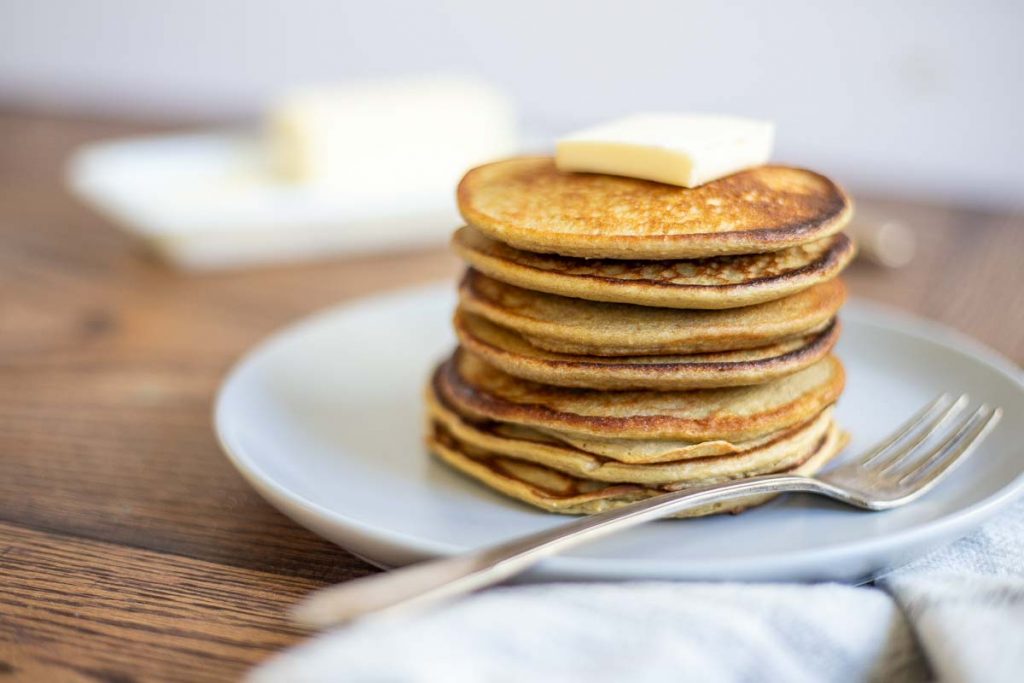 banana protein pancakes stacked up on a gray plate and topped with a pat of butter. The plate sits on a wood table with a white and blue napkin in front