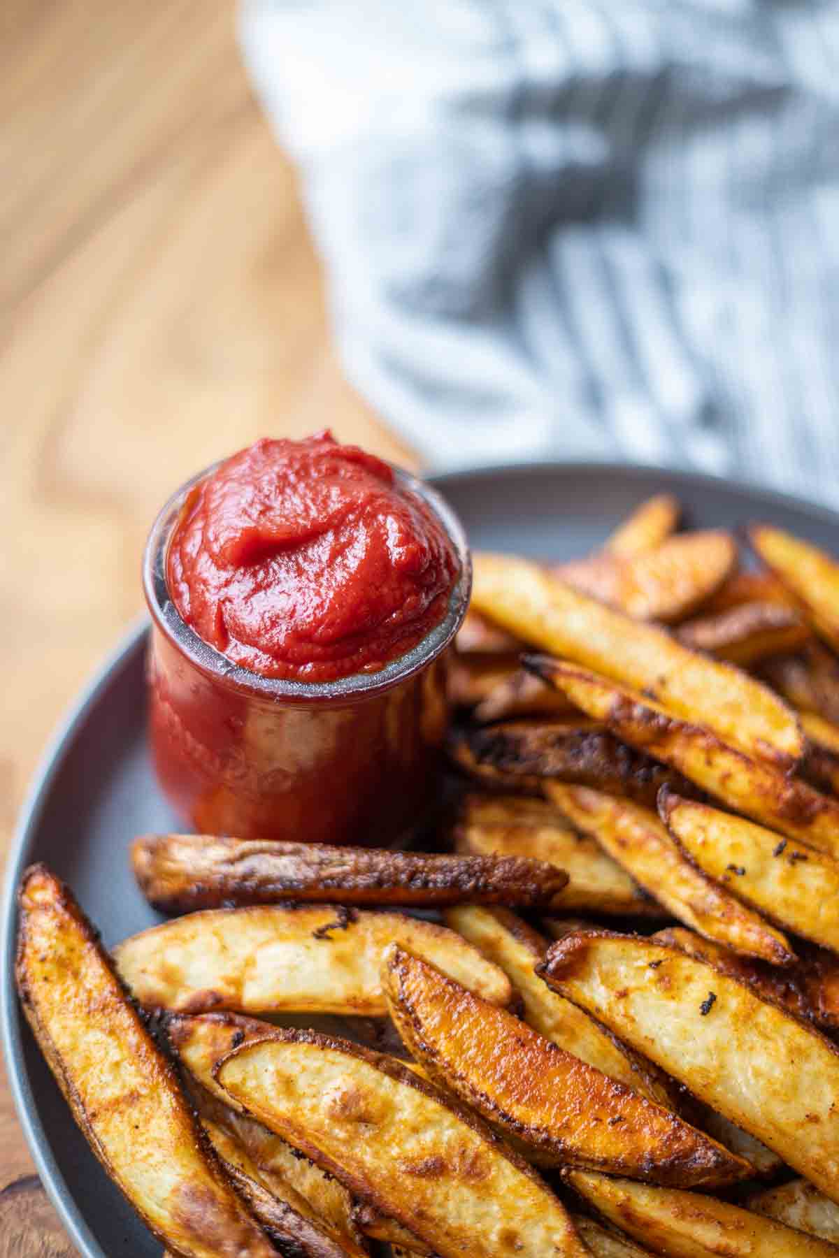 homemade low FODMAP ketchup in a little glass jar on a dark gray plate with roasted potato wedges. A stripped towel is in the background