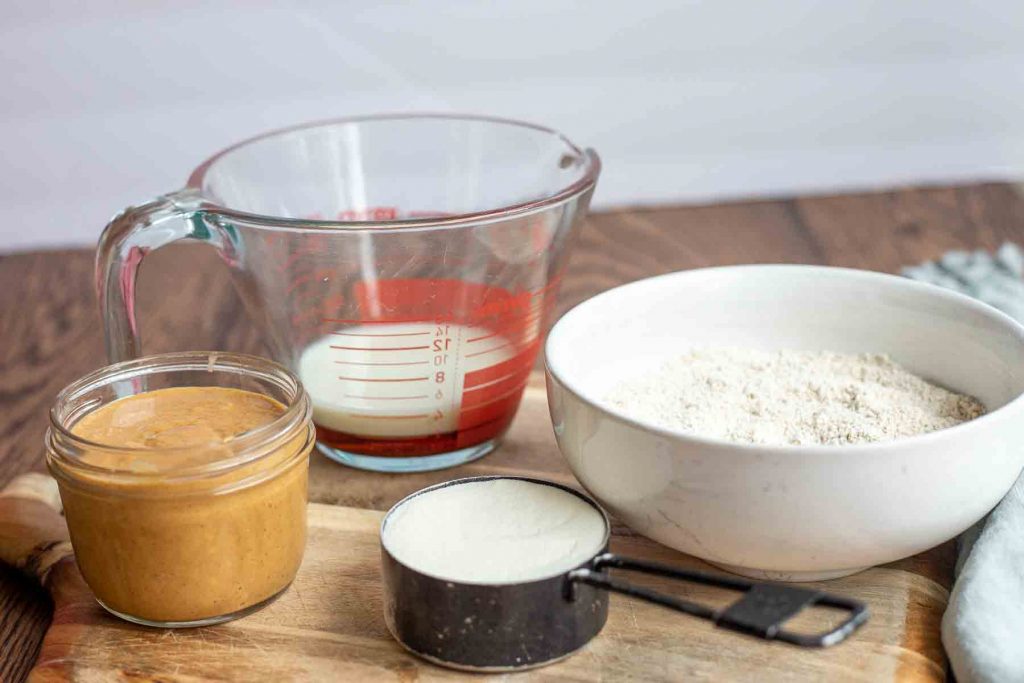 ingredients for bars: peanut butter in clear jar, oat flour in white bowl, collagen in black measuring cup, and maple syrup with oat milk in clear measuring cup on wooden table