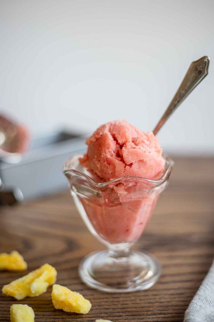 Strawberry pineapple sorbet in clear ice cream dish with spoon onto top of wooden table with chunks of frozen pineapple next to the glass.