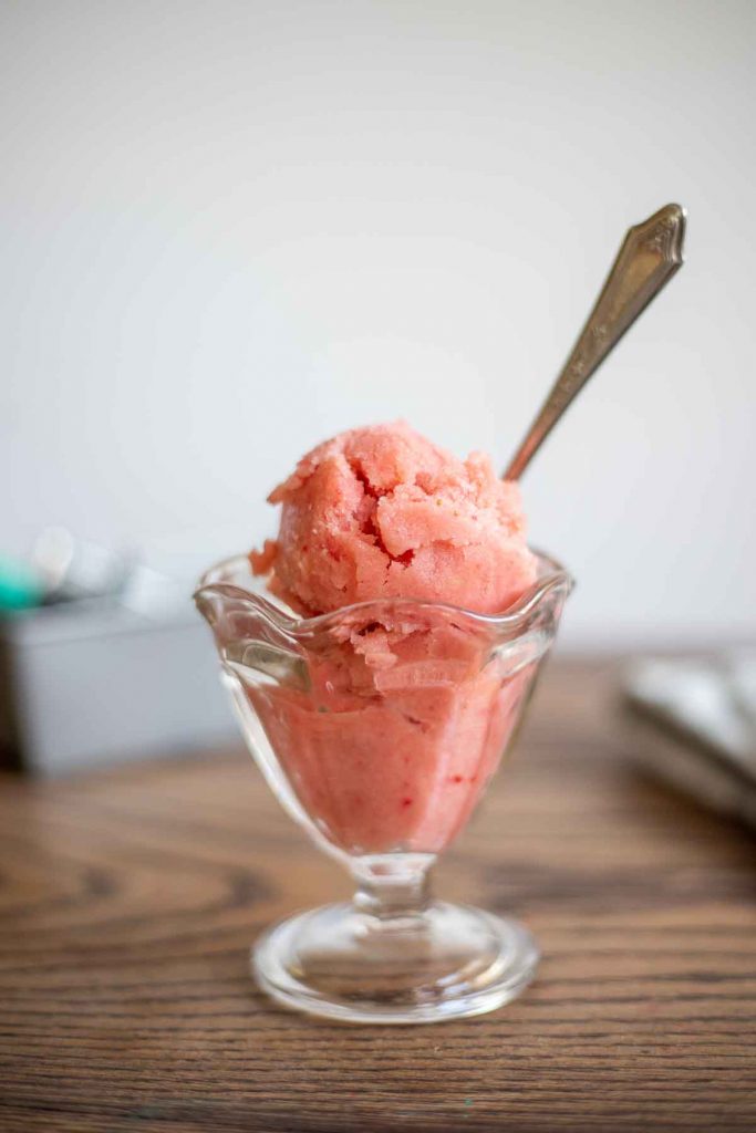 Scoops of strawberry pineapple sorbet in a glass ice cream dish with an antique spoon on a wood table 