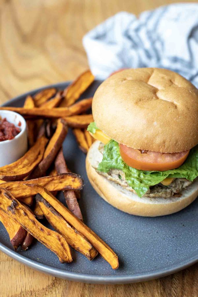 healthy turkey burger loaded with lettuce, tomato, and cheese on a bun with a side of sweet potato fries and a little white jar of ketchup on a gray plate on a wood table