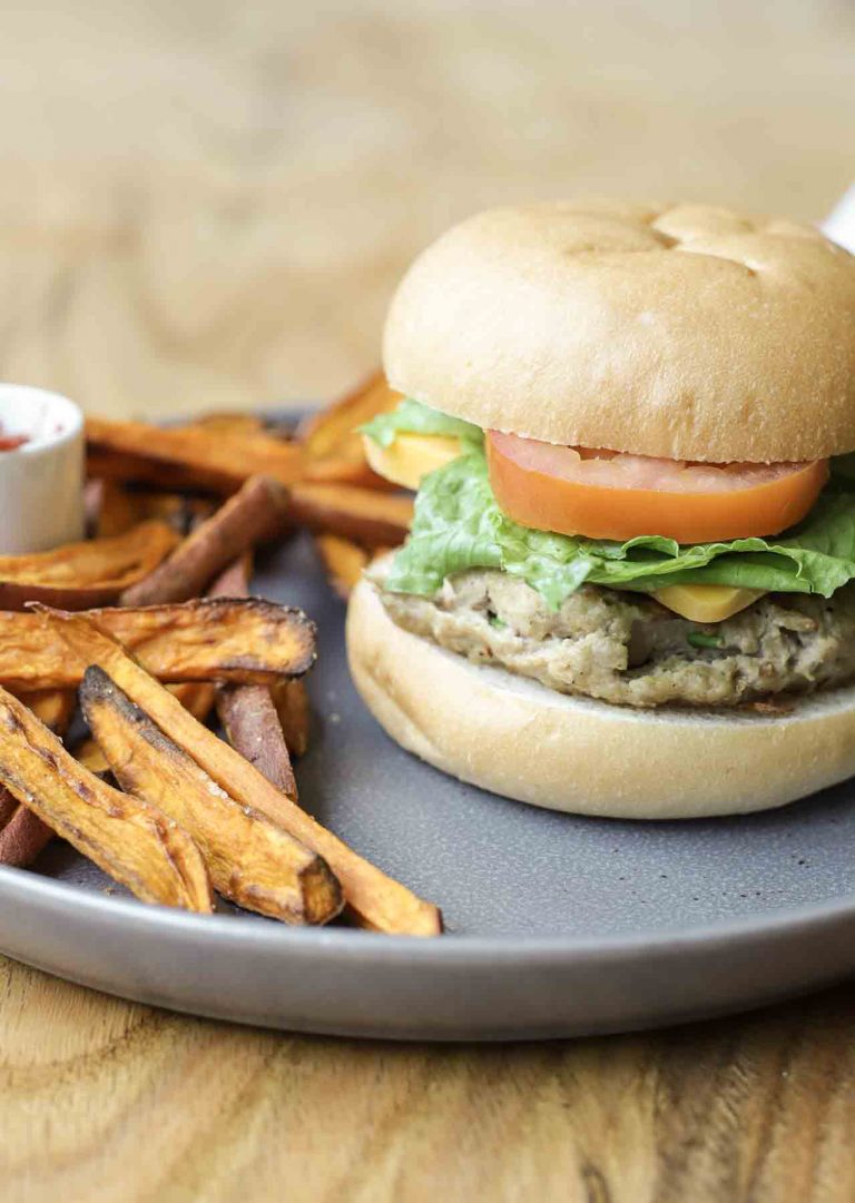 The best healthy turkey burger on a bun with tomato, lettuce, and cheese with a dish of sweet potato fries on a dark gray plate on a wooden table