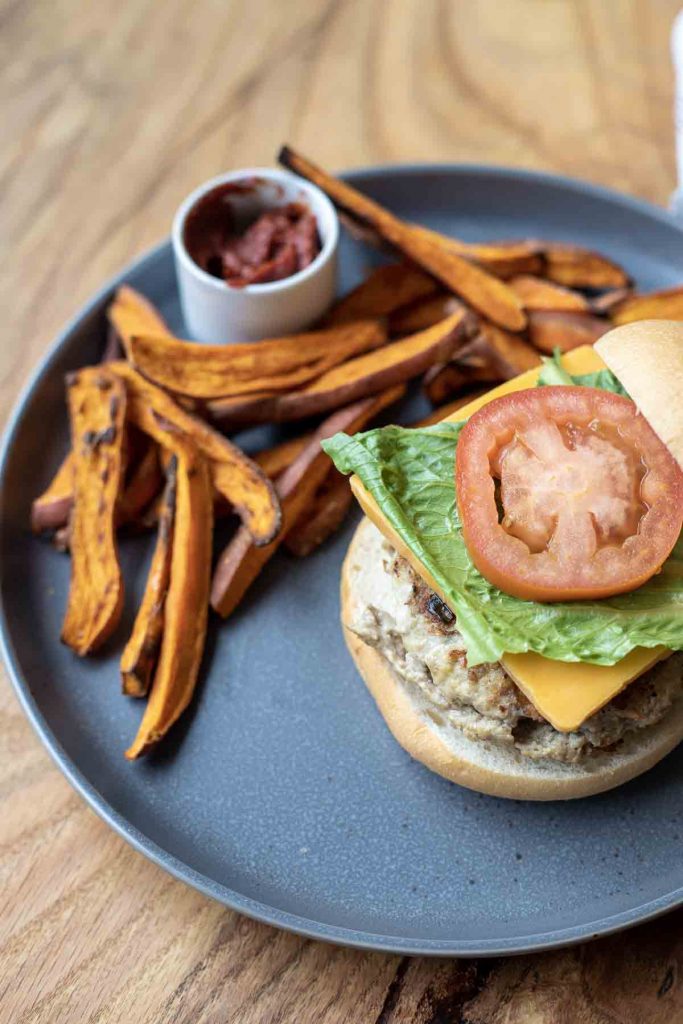 Low FODMAP turkey burger on a bottom bun topped with cheese, lettuce, and tomato with a side of sweet potato fries and ketchup on a dark gray plate on a wood table.
