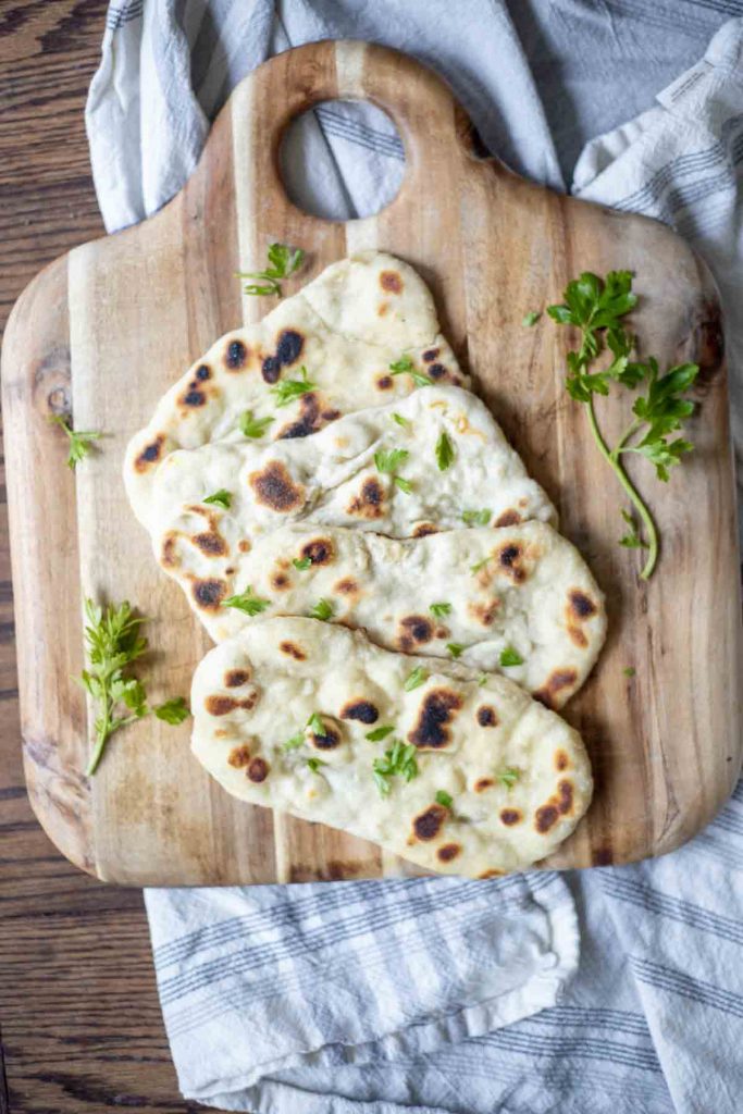 four sourdough flatbreads arranged on a wood cutting board with fresh herbs. The cutting board rests on a blue and white towel