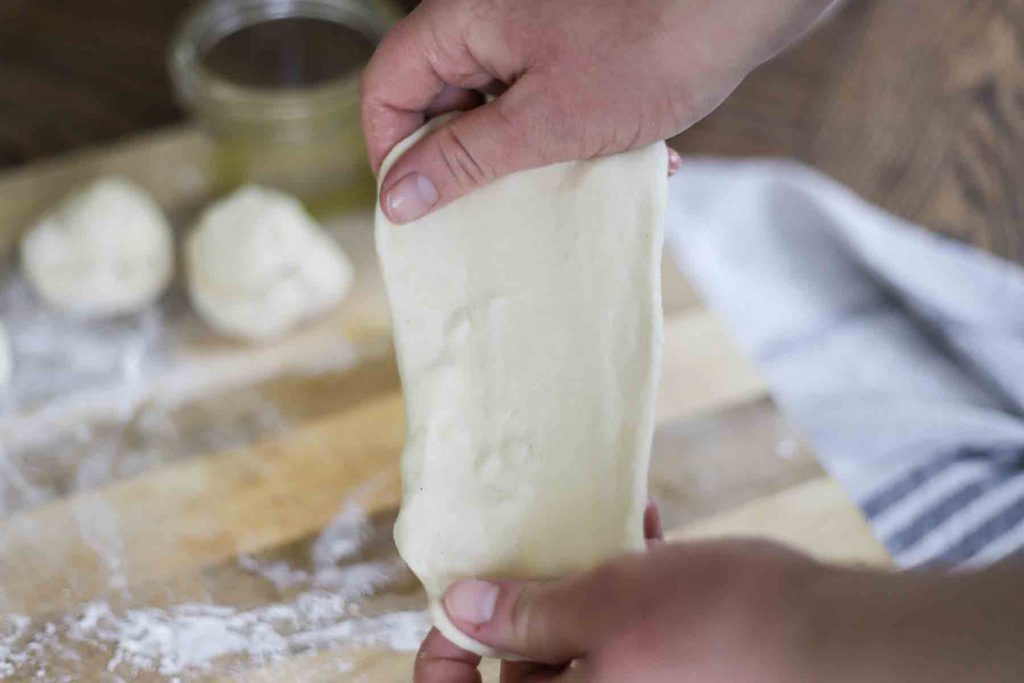 hands stretching out sourdough flatbread dough with a cutting board and dough balls in the background