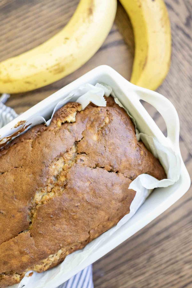 overhead photo of sourdough banana bread in a parchment lined ceramic loaf pan with two ripe bananas