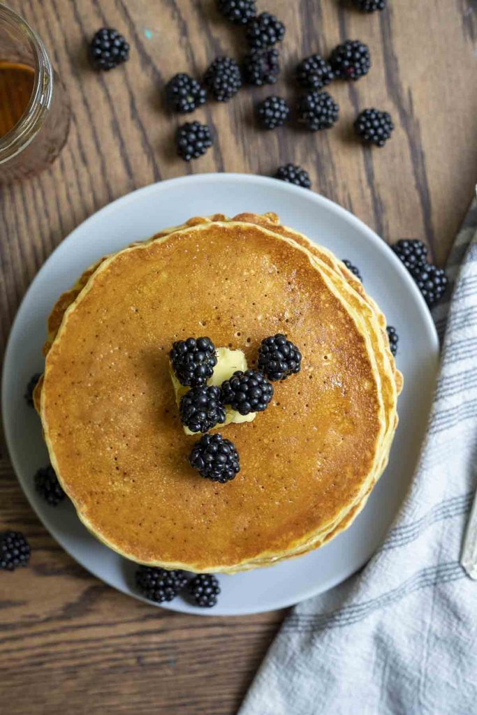 overhead photo of a stack of sourdough pancakes topped with butter and blackberries on a gray plate with more berries scattered around