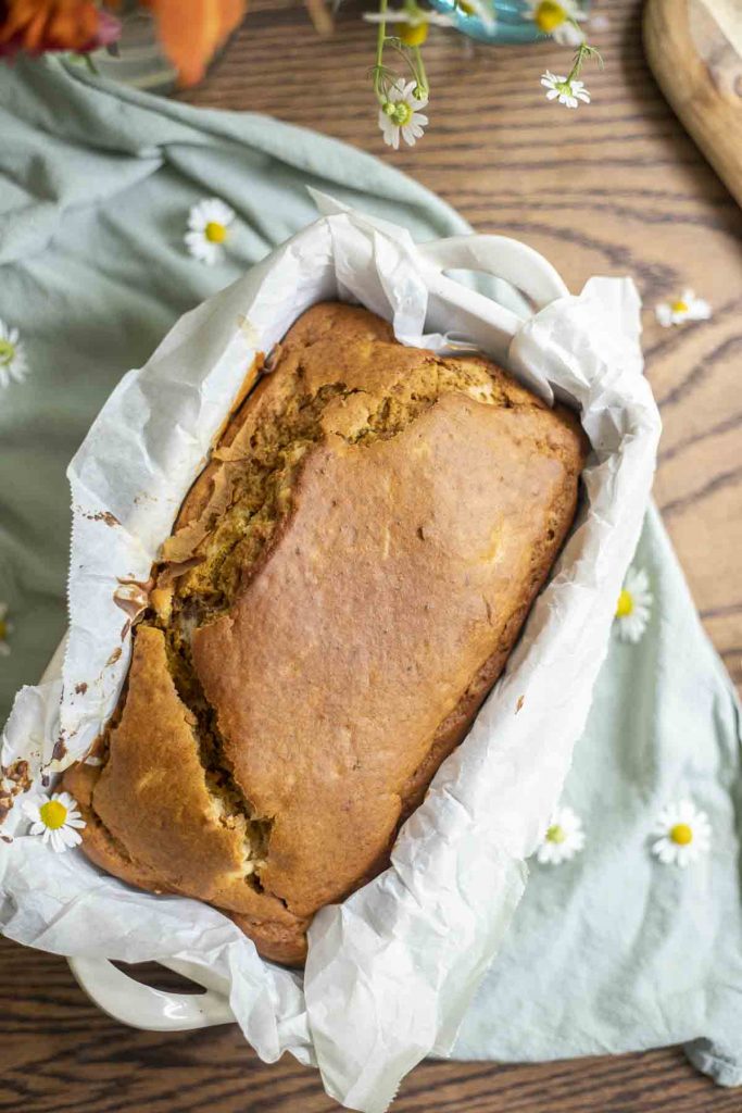 overhead photo of a loaf of sourdough pumpkin bread in a parchment lined loaf pan on a green towel with chamomile flowers spread out around the loaf