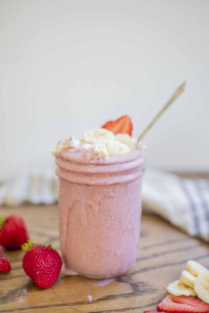 strawberry banansa milkshake in a mason jar topped with whipped cream, sliced bananas and strawberries. the glass sits on wooden table with strawberries and sliced bananas surrounding the glass