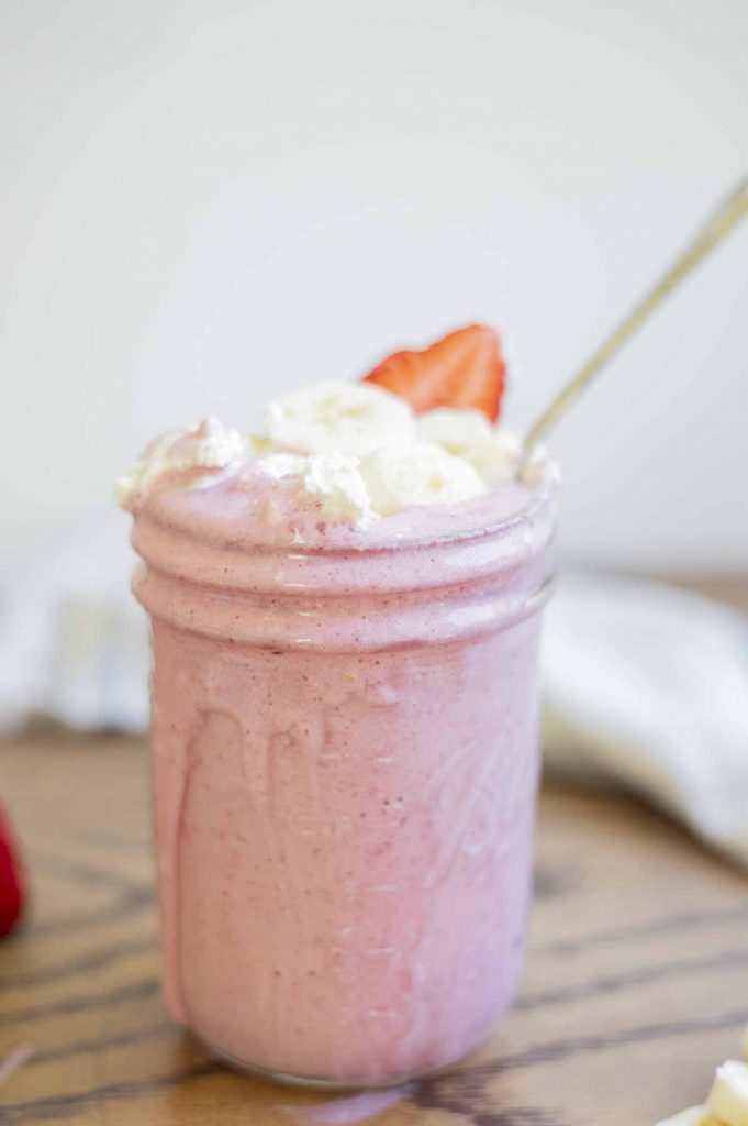 strawberry banana milkshake in a mason jar with a spoon. The milkshake is topped with shipped cream, sliced bananas and strawberries. A cream colored towel is in the background