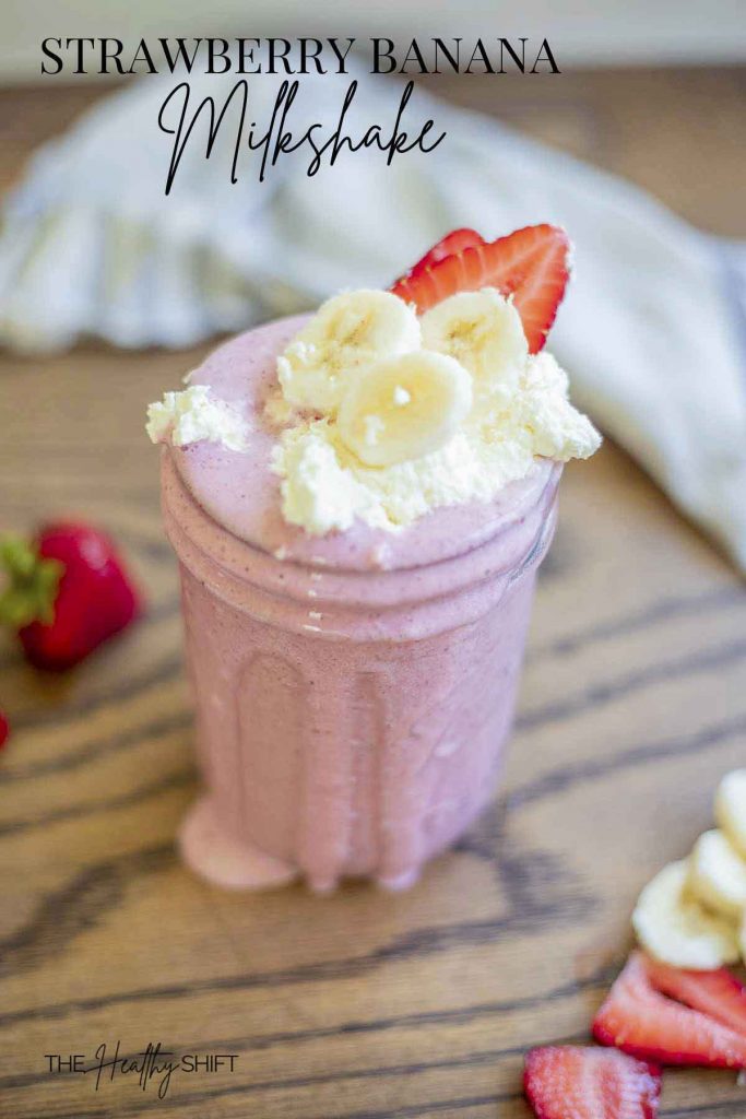strawberry banana milk shake topped with whipped cream, sliced bananas, and sliced strawberries in a mason jar on a wood table surrounded by strawberries and a towel in the background