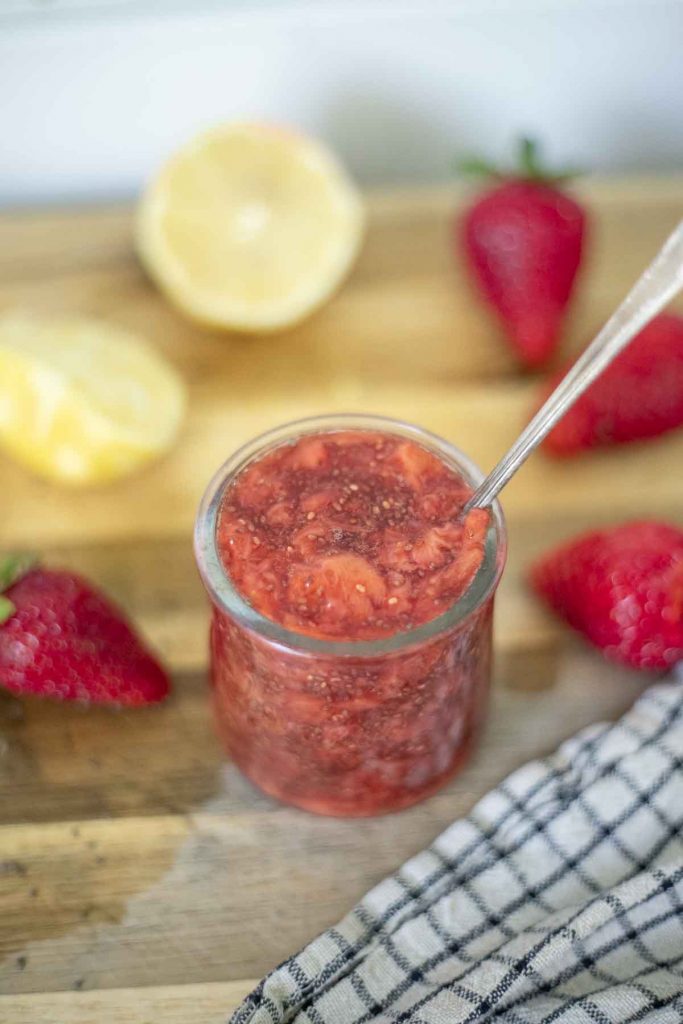 strawberry chia jam in a small glass jar with a spoon on a wood cutting board with strawberries and lemon around the jar