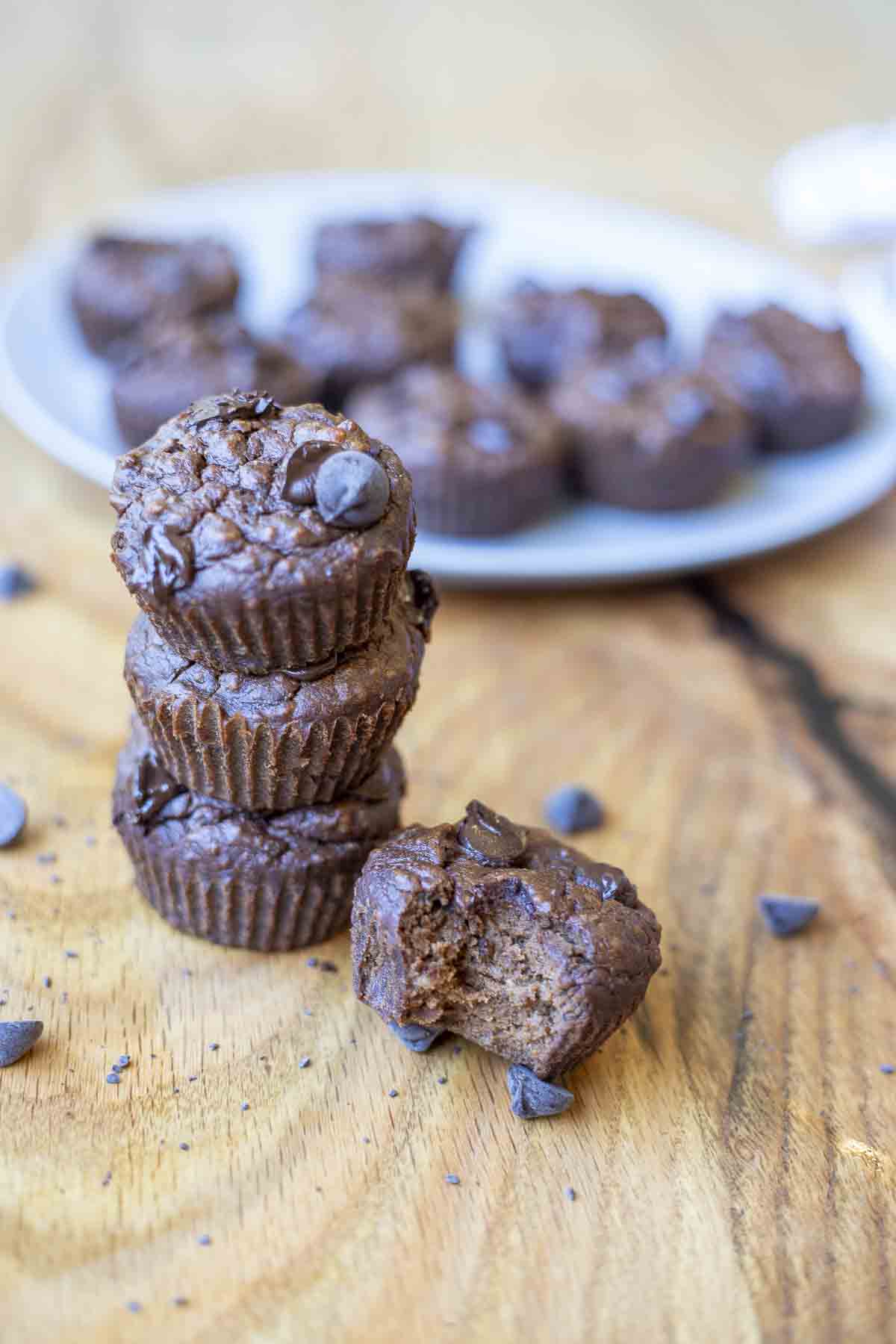 three healthy chocolate muffins stacked up. A fourth muffin is to the right with a bite out. Chocolate chips surround the muffins and there is another pate of muffins in the background